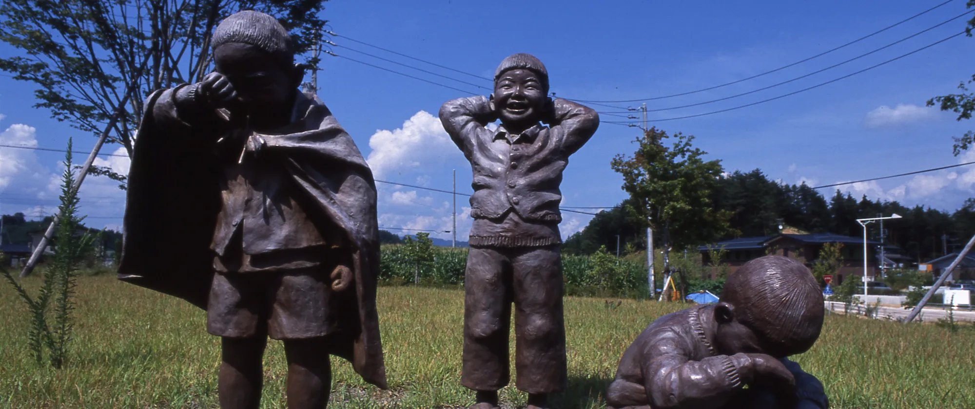 Bronze sculptures of three children on a grassy lawn: one covering his face, one laughing, and one crouching