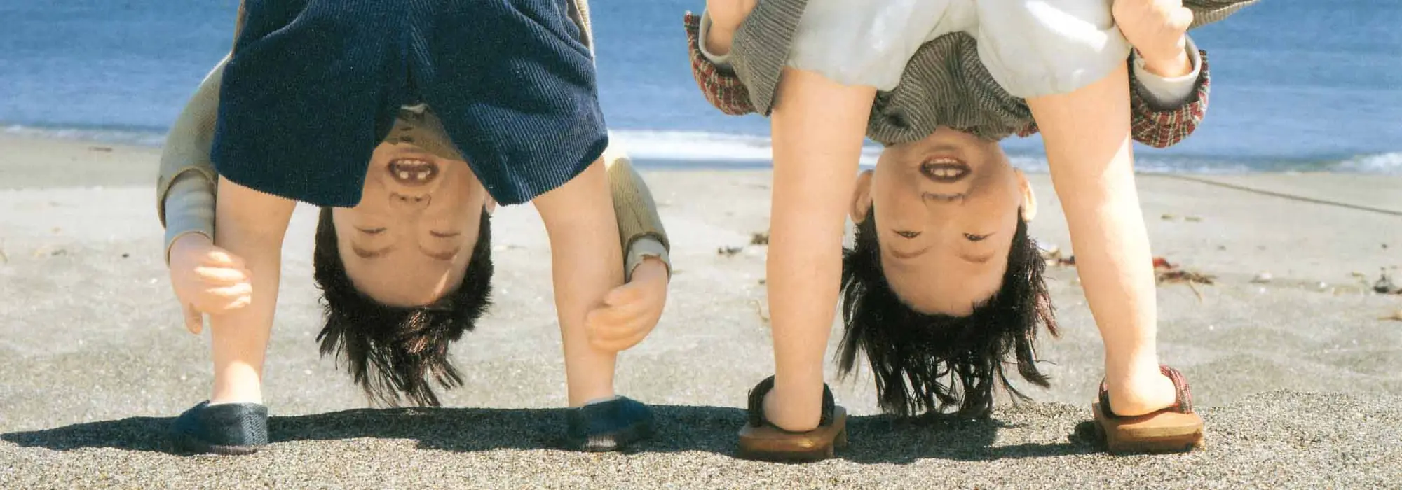 Dolls of two children on a beach, peeking upside down between their legs (the work “Peeking Between the Legs (Matanozoki)”)