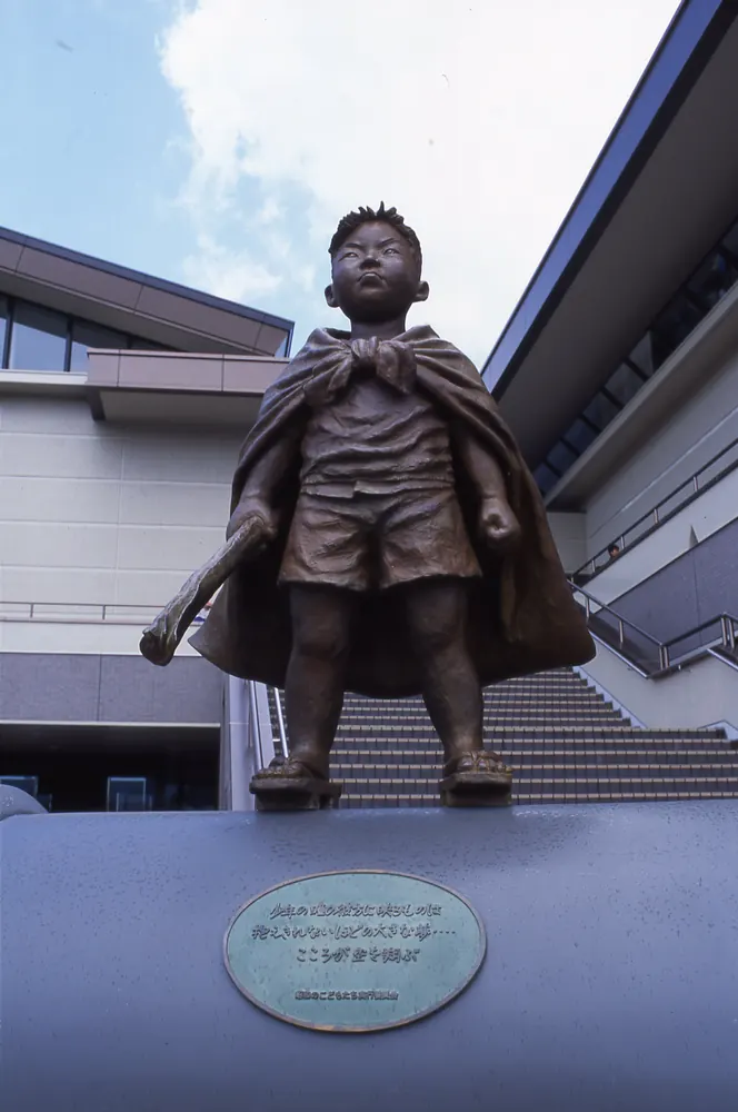 The bronze sculpture “Little Rascal (Wanpaku)”: a boy in a cape standing on a pedestal in front of a building