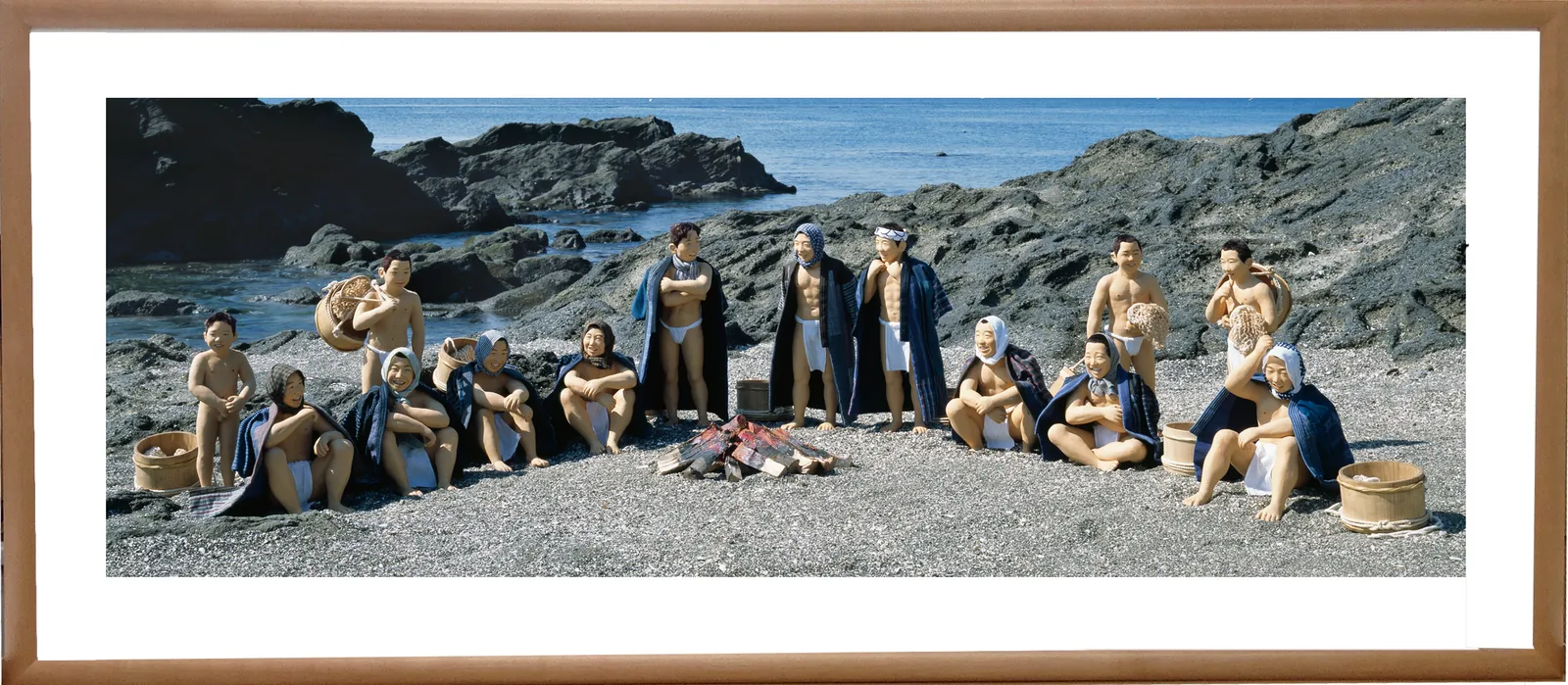 A panorama photo panel in a wooden frame: dolls of male and female divers resting around a bonfire on a rocky shore (the work “The Diver's Rest (Ama no Hitoyasumi)”)