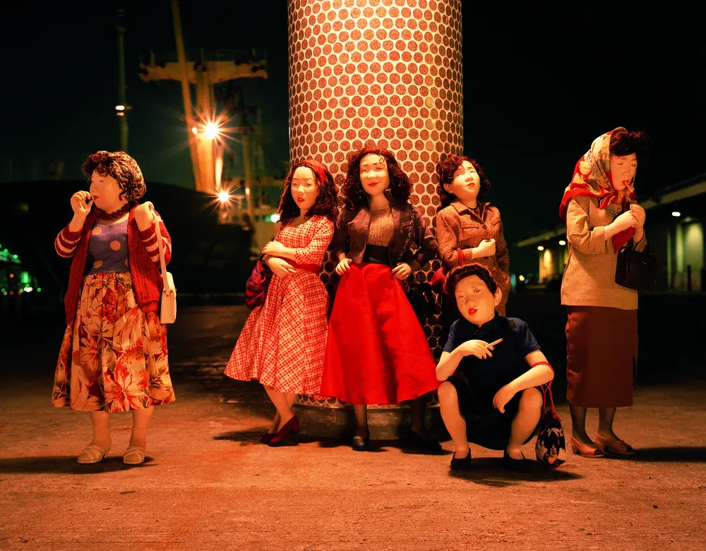 Dolls of women in colorful clothes standing around a lit column at a harbor at night