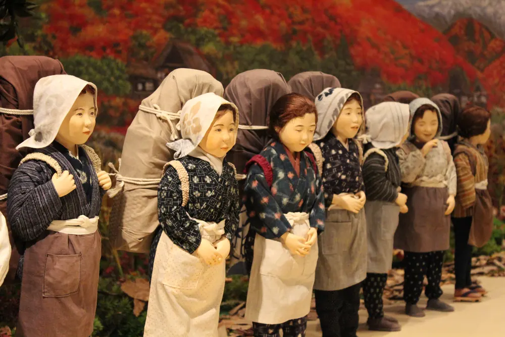 Dolls of peddler women with large loads on their backs, lined up before a painted backdrop of autumn mountains