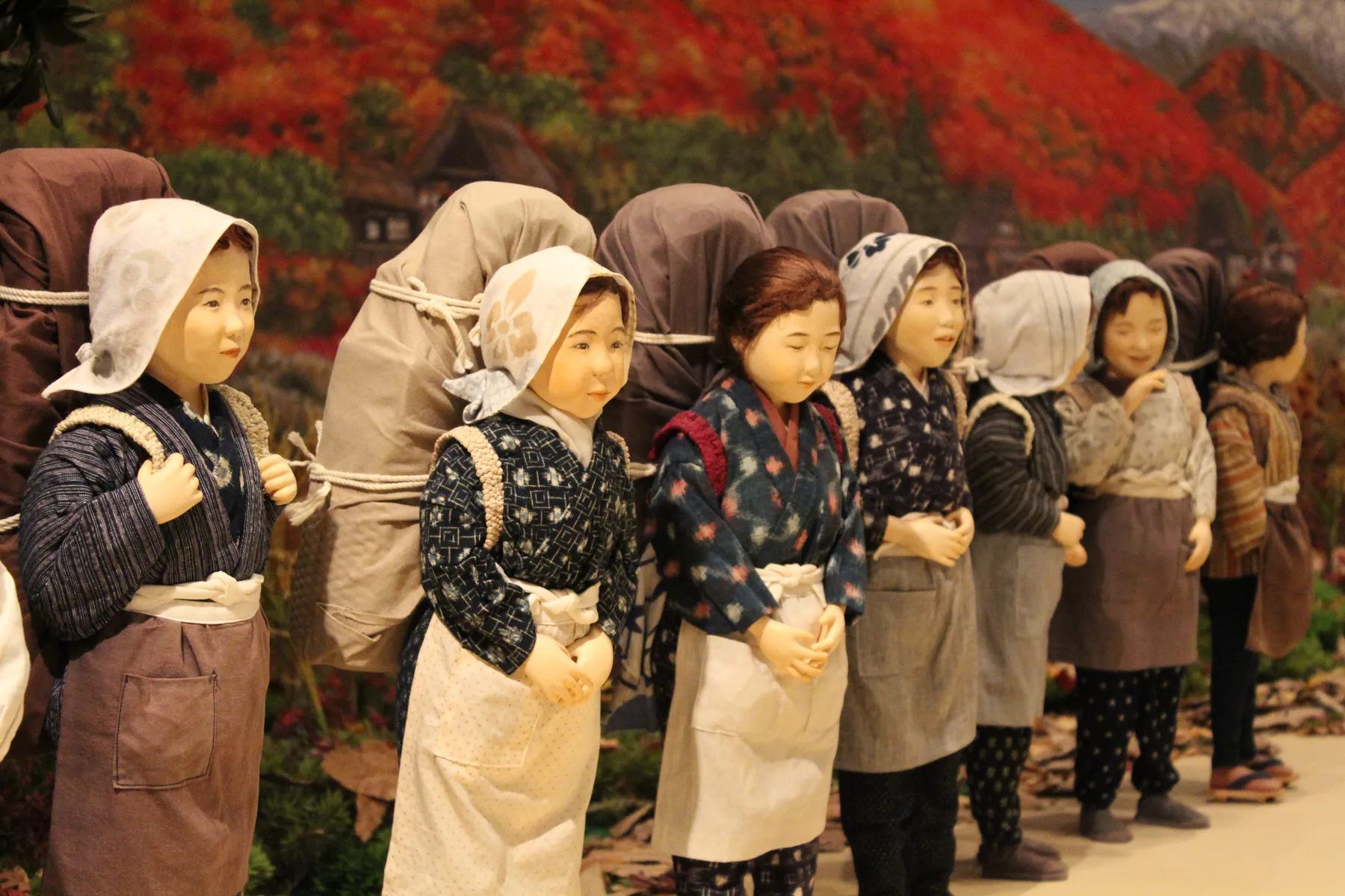 以红叶山景为背景、背着大包袱排成一列的女性人偶（作品《行商的人群》）