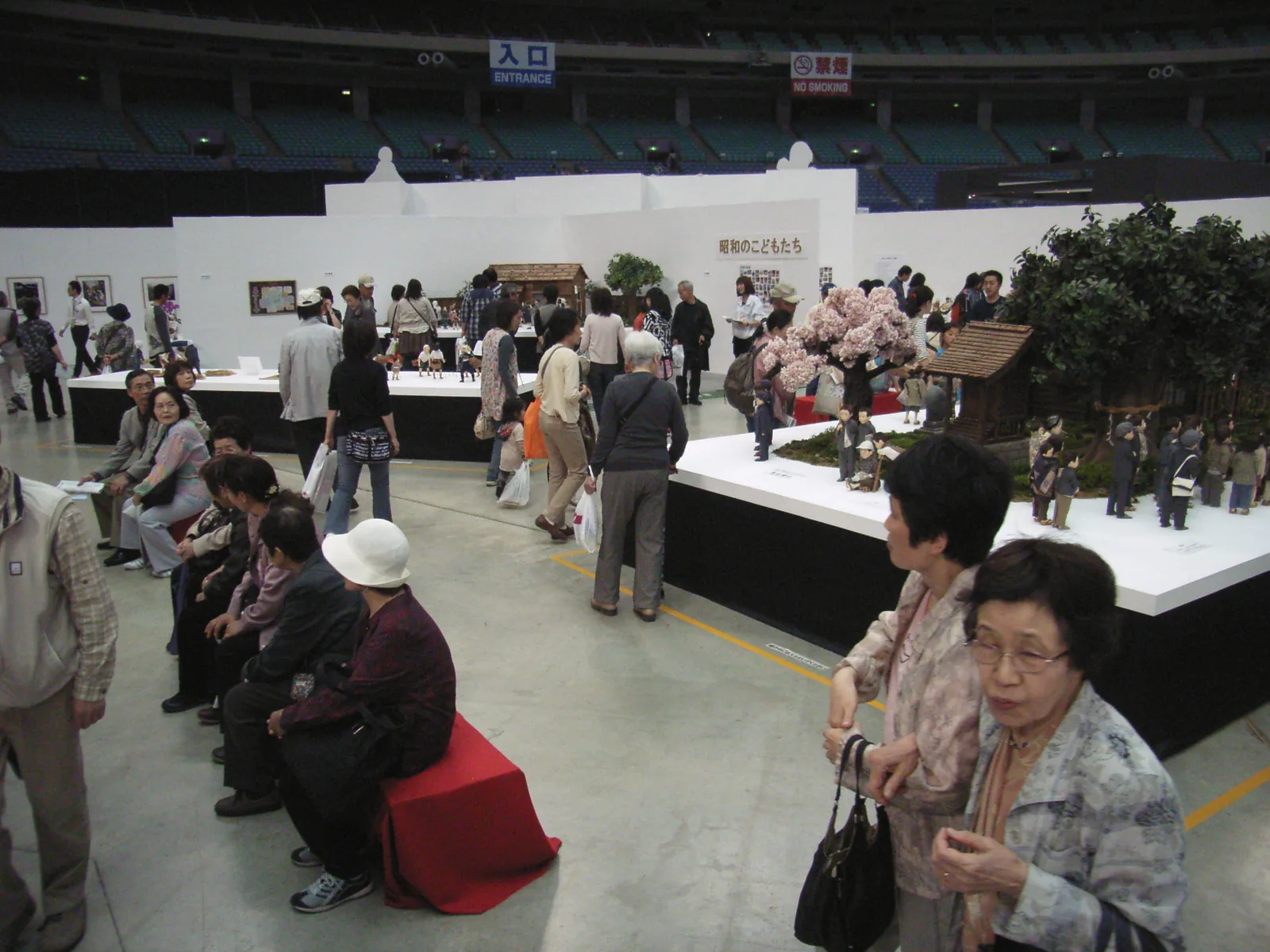広い会場に並ぶ展示台と、桜の木のジオラマ。ベンチで休む人や、展示を見て歩く大勢の来場者