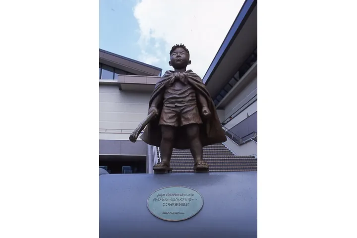 建物の前の台座の上に立つ、マントをはおった男の子のブロンズ像