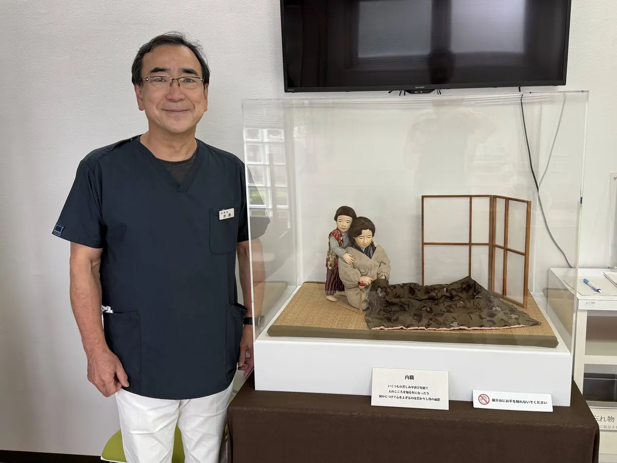 院内の台に置いたアクリルケースの中の作品「内職」の人形と、そのとなりに立つ渡辺医院の理事長