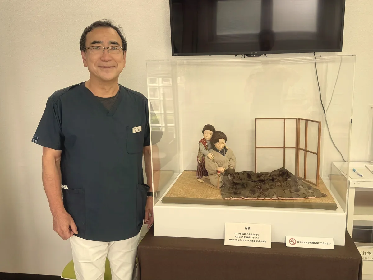 의원 안의 받침대에 놓인 아크릴 케이스 속 작품 〈집안 부업〉의 인형과, 그 옆에 선 와타나베 의원의 이사장