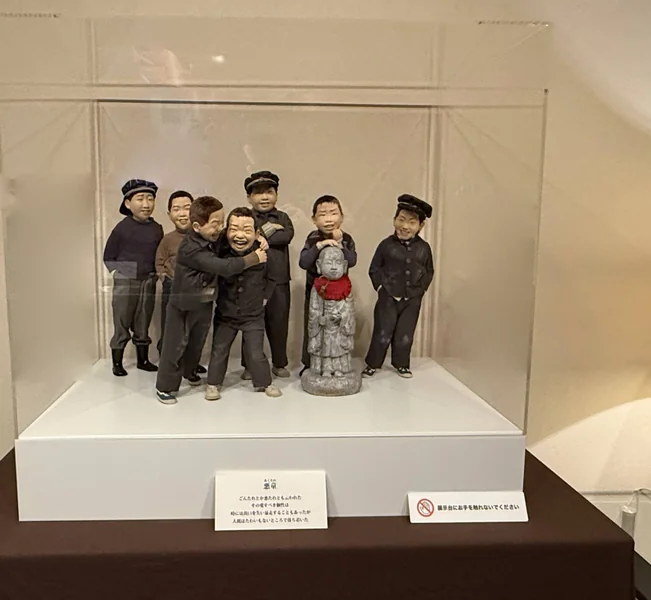 茶色の布を掛けた台の上のアクリルケースと、中の作品「悪童」の人形（肩を組んで笑うこどもたちと、お地蔵さま）。手前に作品の札