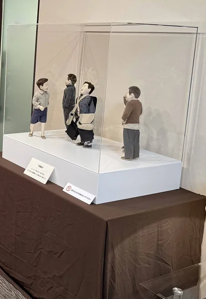 茶色の布を掛けた台の上のアクリルケースと、中の作品「学校帰り」の人形（かばんを掛けたこどもたち）