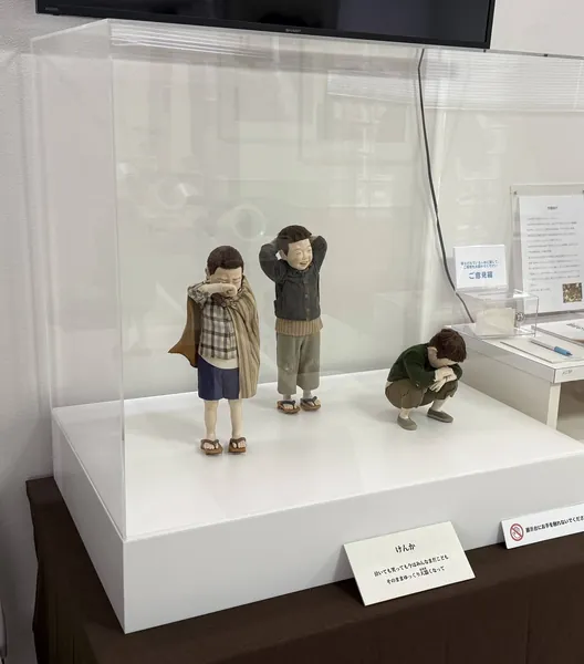 施設の壁ぎわの台に置いたアクリルケースと、中の作品「けんか」の人形（泣く子と笑う子と、うずくまる子）。手前に作品の札