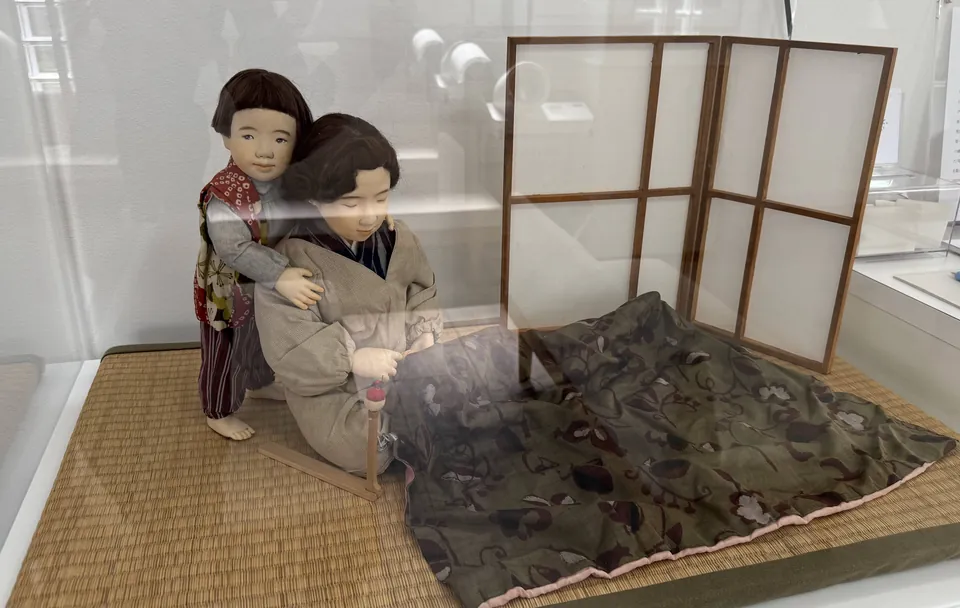 アクリルケースの中の作品「内職」の人形。畳の上で布を広げて手仕事をする母と、その背中に抱きつくこども。うしろに屏風