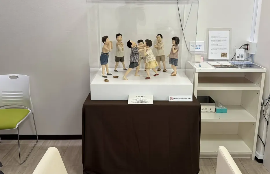 施設の壁ぎわの台に置いたアクリルケースと、中の作品「とっくみあい」の人形。横にいすと棚