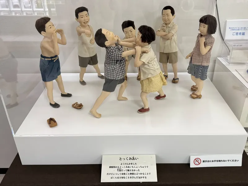 アクリルケースの中で、取っ組み合いをするふたりと、まわりで見ている仲間たちの人形（作品「とっくみあい」）と、作品の札