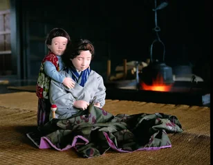 囲炉裏の火のそばで、ひざの上に布を広げて手仕事をする母と、その背中に抱きつくこどもの人形