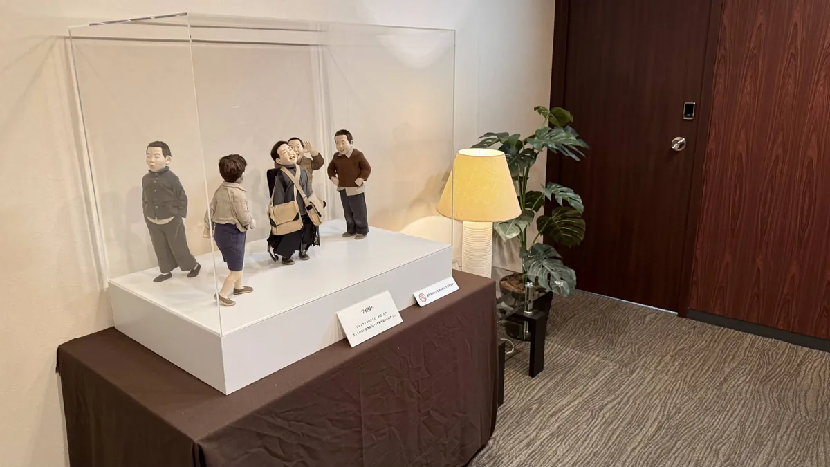 施設の廊下の台に置いたアクリルケースと、中の作品「学校帰り」の5人のこどもの人形。横にスタンドと観葉植物