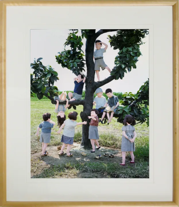 木の額に白いマットで入れた縦の写真パネルのイメージ。大きな木に登ったり、木の下に集まったりするこどもたちの人形の写真（作品「僕らの木」）