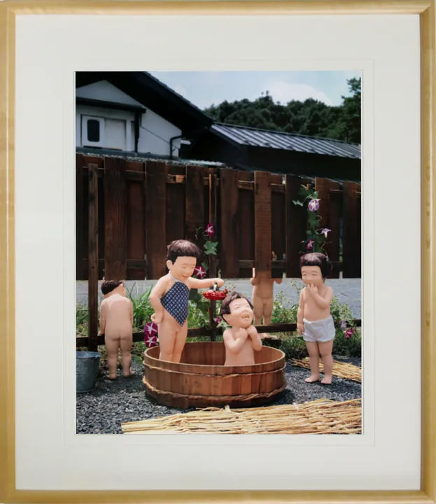 木の額に白いマットで入れた縦の写真パネルのイメージ。板塀の前のたらいで行水をするこどもたちの人形の写真（作品「行水」）