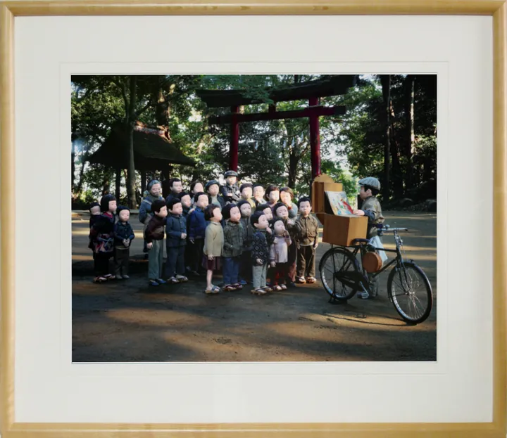 木の額に白いマットで入れた写真パネルのイメージ。神社の境内で、自転車の紙芝居屋さんを囲むおおぜいのこどもたちの人形の写真（作品「紙芝居」）