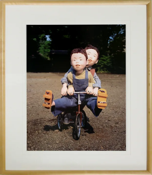 木の額に白いマットで入れた縦の写真パネルのイメージ。三輪車にふたりで乗る男の子たちの人形の写真（作品「三輪車」）