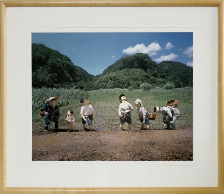 木の額に白いマットで入れた写真パネルのイメージ。山あいの田んぼで、苗を植えたり空を見上げたりするこどもたちと大人の人形の写真（作品「田植え手伝い」）