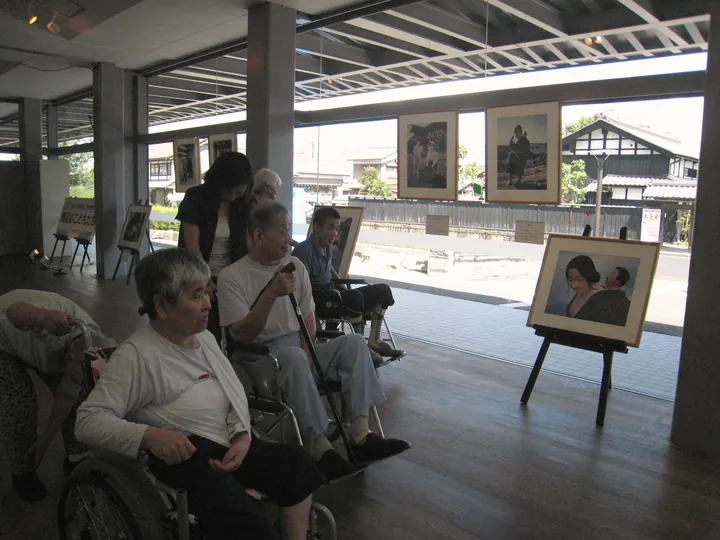 ガラス張りの通路に三脚で並べた写真パネルと、いすに座って見る人たち