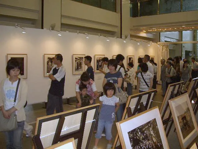 展示室の壁と三脚に並べた写真パネルを見る、大勢の人たち