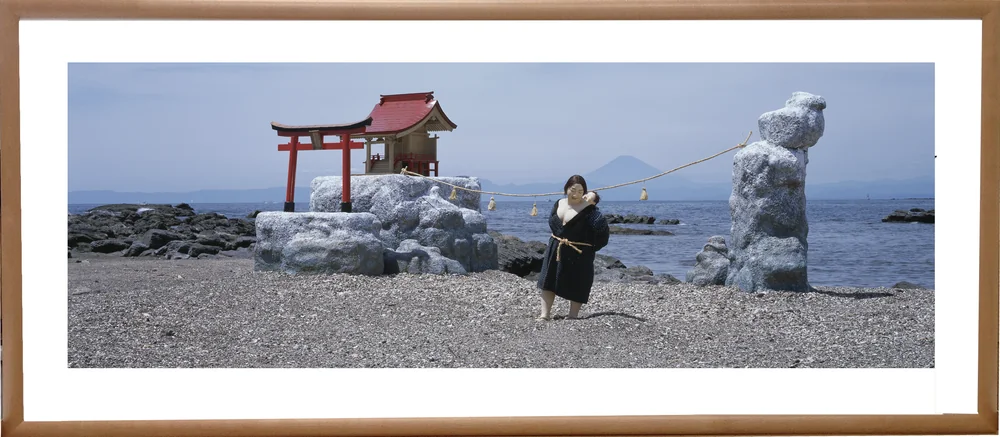 木の額に入れた横長の写真パネルのイメージ。浜辺の鳥居とほこらを背に、赤ちゃんを背負って立つ、縄の帯をしめた着物の女性の人形（作品「永遠の祈り」）
