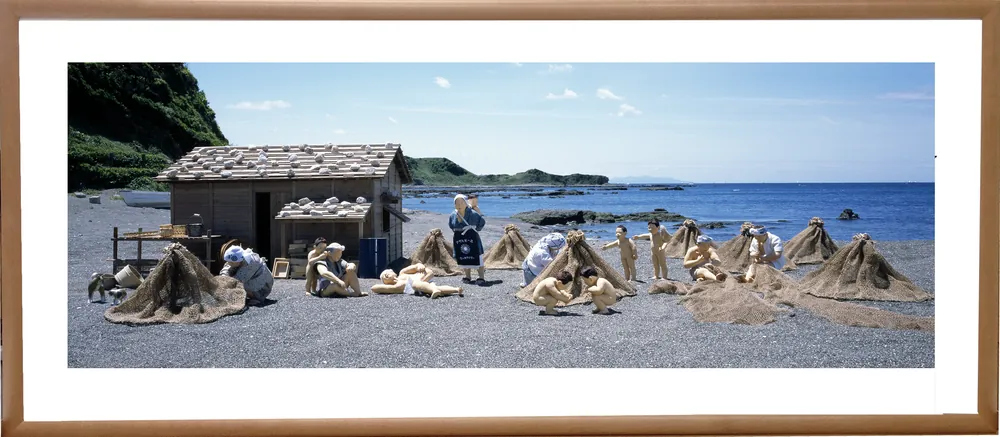 木の額に入れた横長の写真パネルのイメージ。浜辺に網を干した小屋のまわりで過ごす、海の人々とこどもたちの人形（作品「浜の午後」）